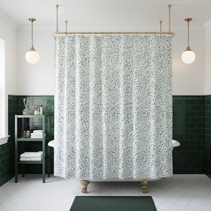 May include: A bathroom with a white and green color scheme. A shower curtain with a leaf pattern hangs from a brass rod. The walls are white with green tile accents. A green rug sits on the floor. Two globe pendant lights hang from the ceiling.