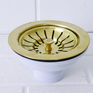 May include: A gold-colored sink strainer with a white plastic base. The strainer has a circular design with a small knob in the center.