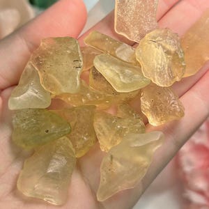 Puede incluir: Un puñado de piezas de vidrio de mar translúcidas, de forma irregular y de tono amarillo. Los fragmentos de vidrio varían en tamaño y tienen un aspecto liso y esmerilado. Las piezas se sostienen en una mano, con un fondo suave.