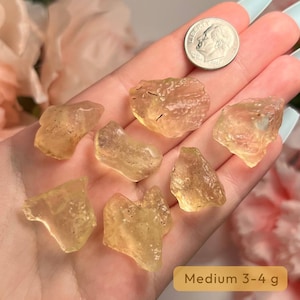 Puede incluir: Siete gemas en bruto de color amarillo dorado, con formas irregulares. Una moneda de plata se utiliza para comparar el tama&ntilde;o. El texto "Medium 3-4 g" aparece sobre un fondo beige. Las piedras pesan entre 3 y 4 gramos.