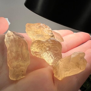 Puede incluir: Cuatro piedras preciosas en bruto transl&uacute;cidas de color &aacute;mbar se sostienen en una mano. Las piedras tienen formas irregulares y una textura rugosa. La iluminaci&oacute;n resalta el color y la claridad de las piedras, lo que sugiere que son cristales o minerales.