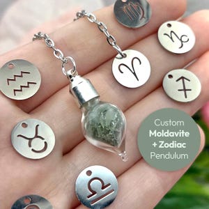 May include: A silver chain pendulum with a glass vial filled with green moldavite crystals. The pendulum is attached to a silver charm with a zodiac symbol. The image also shows other zodiac charms in the background.