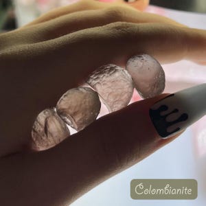 May include: A close-up of a hand holding several small, translucent, grey stones. The stones have a rough, textured surface. The text "Colombianite" is visible in the lower right corner of the image.