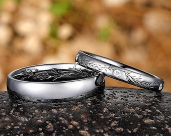 Può includere: Due fedi nuziali d'argento. Un anello ha un disegno a foglie all'interno e un esterno lucido. L'altro anello ha un disegno a foglie con piccole pietre trasparenti. Gli anelli sono su una superficie scura e strutturata.