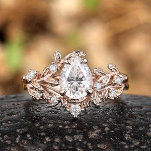 May include: A pear-shaped diamond engagement ring set in rose gold with a delicate floral design. The ring features a pear-shaped center stone surrounded by smaller diamonds and intricate leaf details.