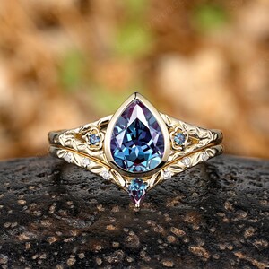 May include: A gold engagement ring set with a pear-shaped, color-changing alexandrite gemstone. The ring is accented with small blue gemstones and delicate floral detailing. The band is also gold and features a matching floral design.
