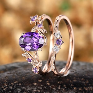 May include: A rose gold ring set featuring an oval-cut purple gemstone and smaller purple and white accent stones. The engagement ring has a floral design, and the wedding band has a delicate pattern. The rings are displayed on a dark, textured surface.