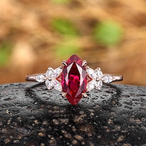 May include: A rose gold ring with a marquise-cut, deep red gemstone as the focal point. The ring is adorned with small, white flower-shaped accents on either side of the central stone. The ring is set against a dark, textured background.