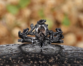 May include: Black engagement ring with a large, clear, oval-shaped gemstone surrounded by smaller black gemstones. The ring band is black and features a delicate, leafy design.