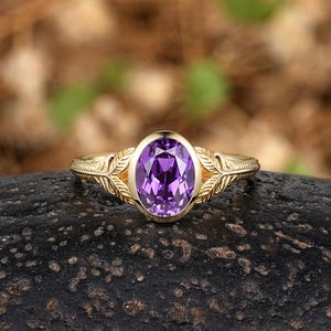 May include: A gold ring featuring a central oval-cut purple amethyst gemstone in a bezel setting. The band is crafted with detailed leaf-like engravings extending from the center stone along the sides, creating a textured and ornate appearance.