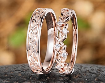 May include: Two rose gold rings. One ring features a leaf pattern, and the other has a floral design with small, clear gemstones. The rings are displayed on a dark, textured surface against a blurred background.