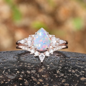 Könnte beinhalten: Ein Verlobungsring mit einem birnenförmigen Opal, der in Roségold gefasst ist, mit einem zarten Band aus kleineren Diamanten. Der Ring liegt auf einer braunen, strukturierten Oberfläche.