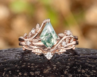 Puede incluir: Anillo de compromiso de oro rosa con una piedra preciosa de ágata verde musgo engastada en un delicado diseño de hojas con pequeños diamantes. El anillo está sobre una superficie marrón oscura.