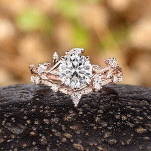 May include: A rose gold engagement ring with a large round center stone and smaller stones set in a delicate vine design.
