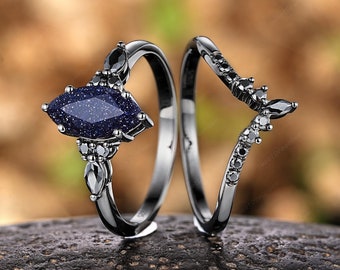 May include: A black ring set with a blue marquise-shaped gemstone and black diamonds. The engagement ring features a larger gemstone and the wedding band has smaller black diamonds.