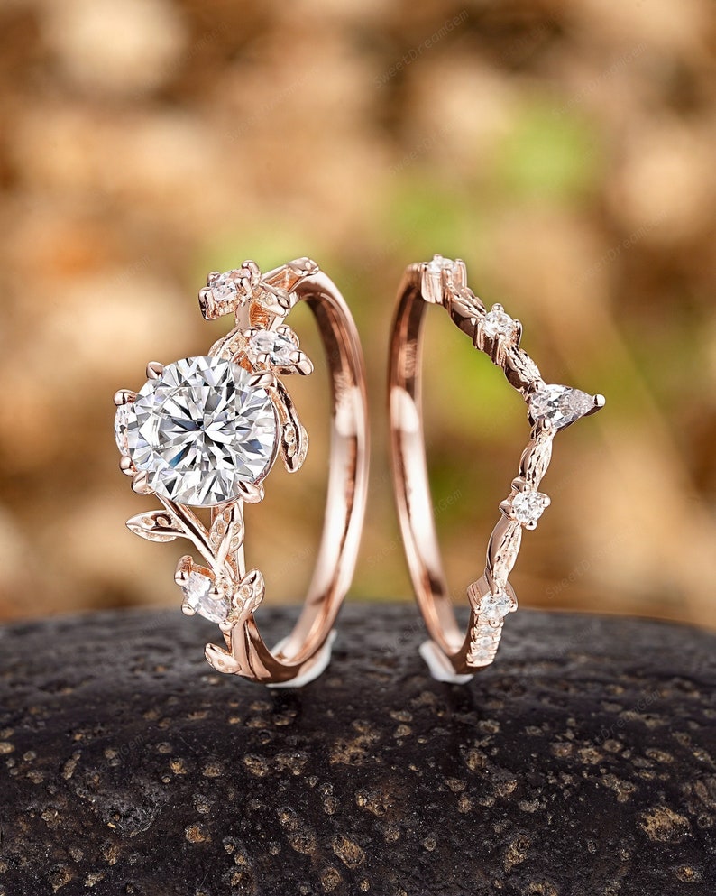 May include: A set of two rose gold engagement rings with delicate floral designs and sparkling diamonds. The larger ring features a round center stone surrounded by smaller diamonds and leaves. The smaller ring has a curved band with a row of diamonds and leaves.