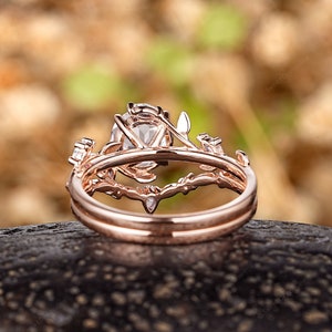 May include: A rose gold engagement ring set with a large oval-shaped center stone and delicate floral details. The ring is set on a dark brown surface.