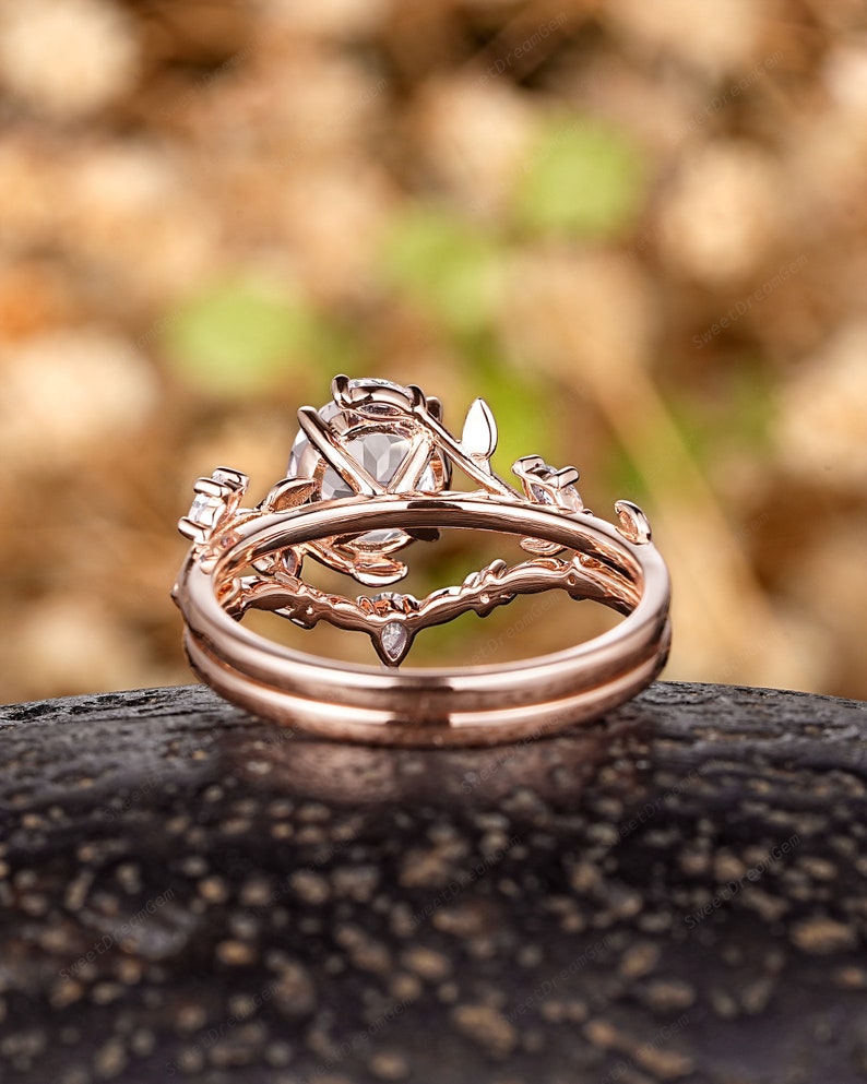 May include: A rose gold engagement ring set with a large oval-shaped center stone and delicate floral details. The ring is set on a dark brown surface.