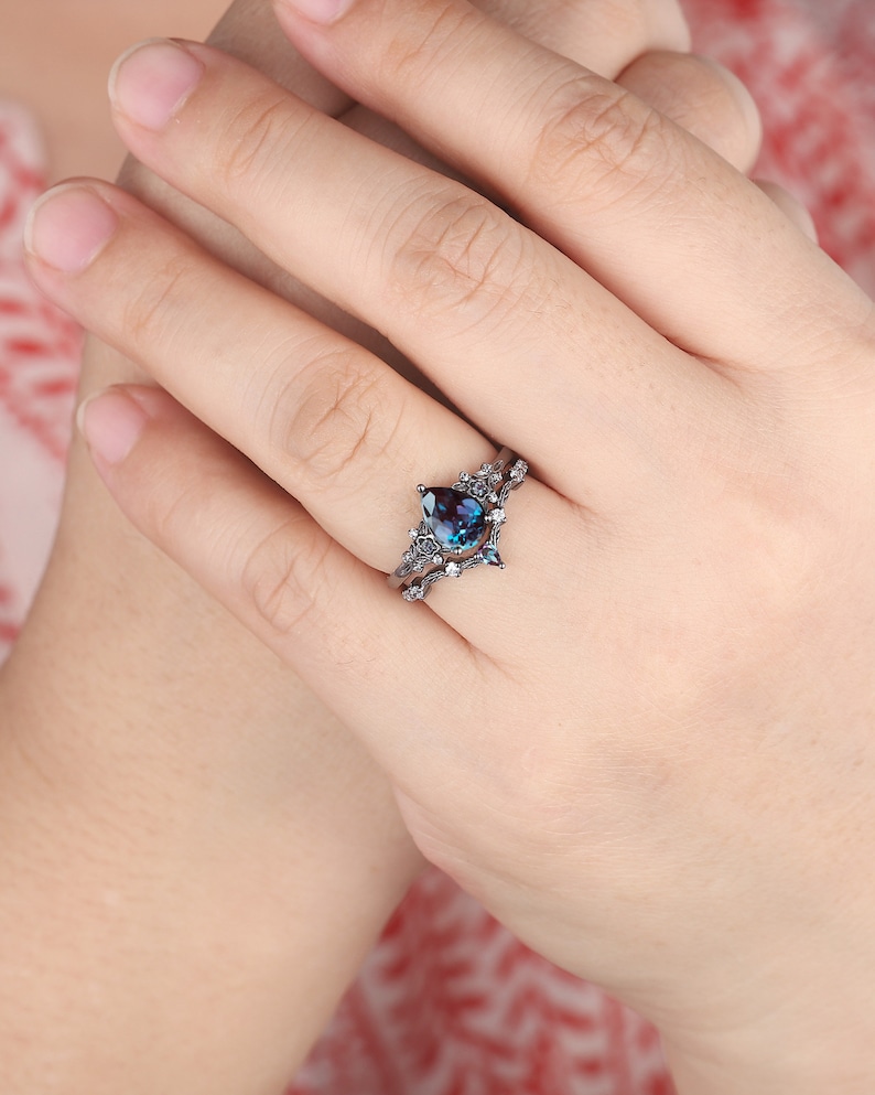 May include: A pear-shaped blue gemstone ring with a black metal setting and a delicate floral design. The ring is set on a white band.