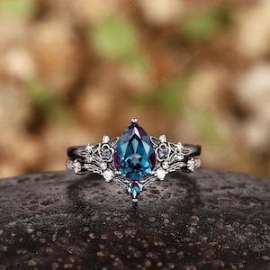 May include: A pear-shaped alexandrite engagement ring set in black rhodium plated sterling silver. The ring features a delicate floral design with smaller white diamonds and a single black diamond.