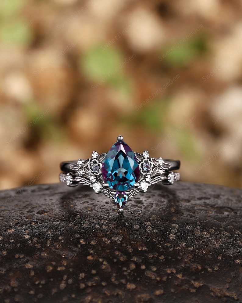 May include: A pear-shaped alexandrite engagement ring set in black rhodium plated sterling silver. The ring features a delicate floral design with smaller white diamonds and a single black diamond.