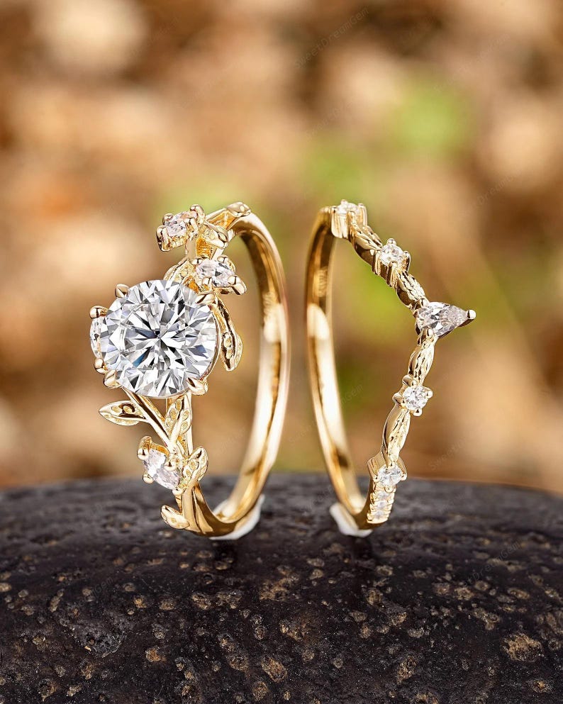 May include: A set of two gold rings with delicate floral designs and sparkling clear stones. The larger ring features a round center stone and smaller stones on the branches. The smaller ring has a curved design with smaller stones.