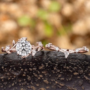 May include: A rose gold engagement ring set with a large round center stone and smaller stones set in a delicate leaf design. The matching wedding band features a similar leaf design with a smaller pear-shaped stone.