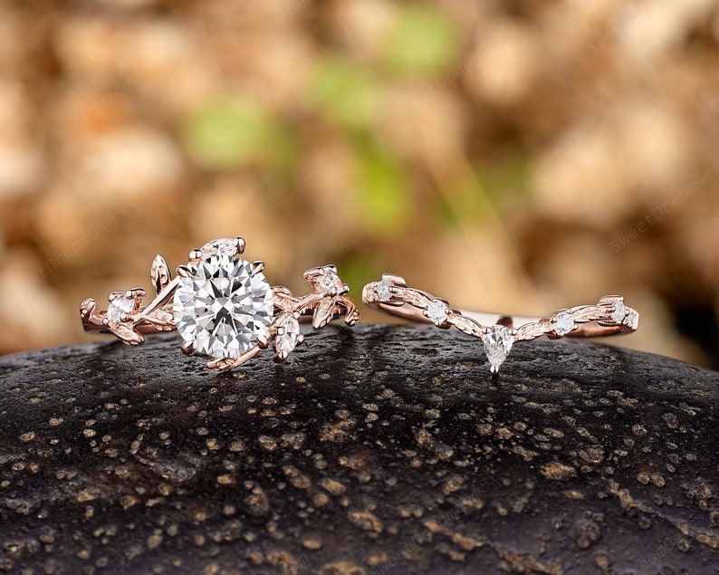 May include: A rose gold engagement ring set with a large round center stone and smaller stones set in a delicate leaf design. The matching wedding band features a similar leaf design with a smaller pear-shaped stone.