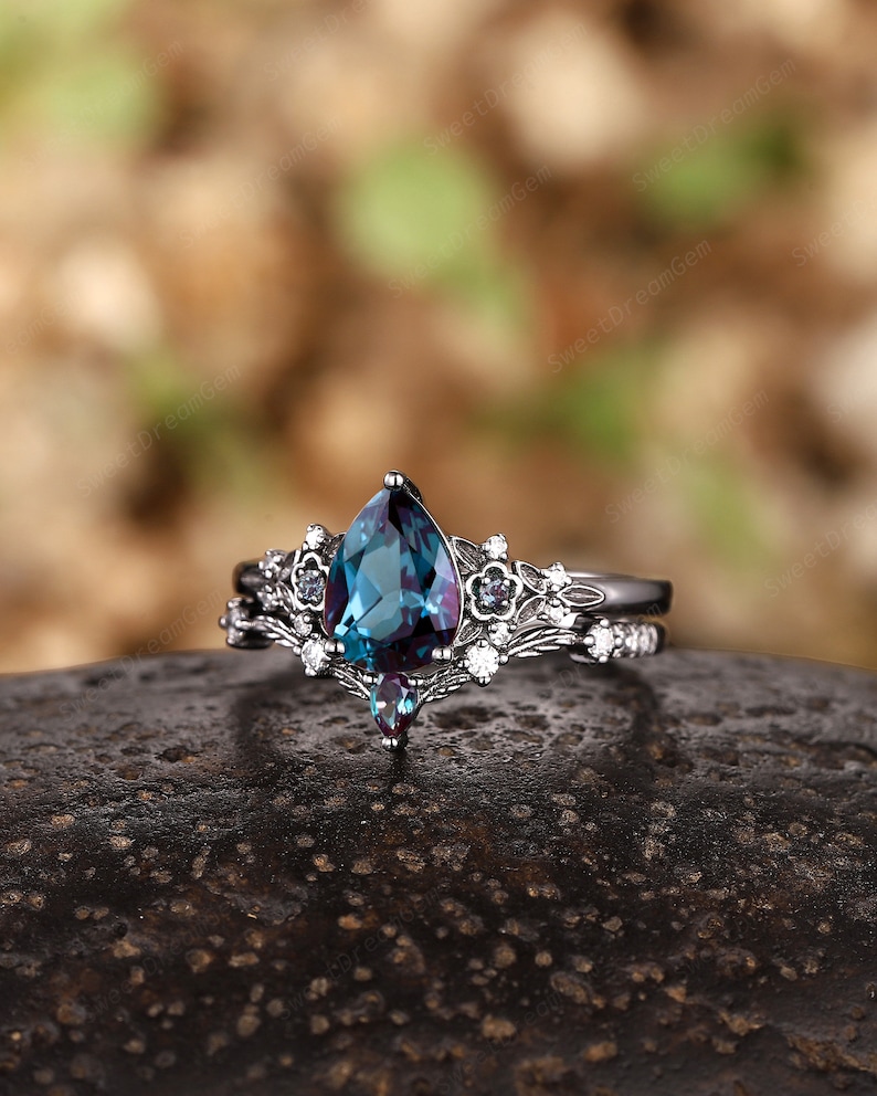 May include: A pear-shaped, blue-green gemstone ring with a black metal band and a delicate floral design. The ring is set with smaller white gemstones and a single, smaller blue-green gemstone.