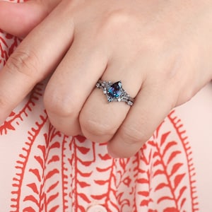 May include: A black ring with a pear-shaped blue gemstone and smaller clear gemstones. The ring is on a finger wearing a white and red patterned shirt.