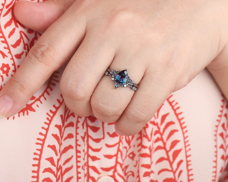 May include: A black ring with a pear-shaped blue gemstone and smaller clear gemstones. The ring is on a finger wearing a white and red patterned shirt.