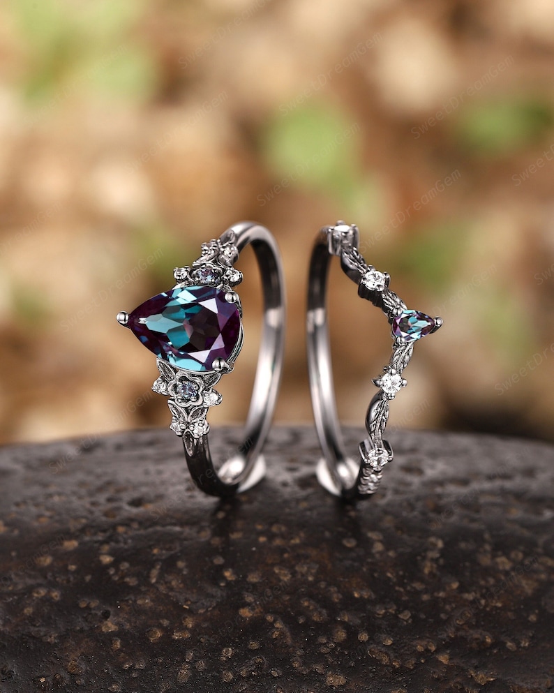 May include: A pair of silver rings featuring a pear-shaped alexandrite gemstone set in the centre of the larger ring. The smaller ring has a delicate floral design with small clear gemstones.