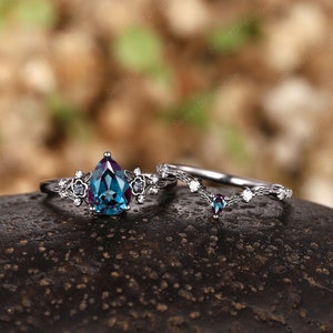 May include: A pear-shaped blue and green gemstone engagement ring with a delicate floral design and a matching wedding band with a V-shaped design.