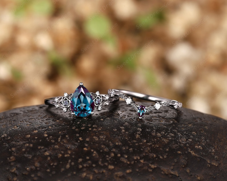 May include: A pear-shaped blue and green gemstone engagement ring with a delicate floral design and a matching wedding band with a V-shaped design.
