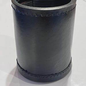 May include: A cylindrical black holder with a smooth, textured surface and visible stitched detailing along the top and bottom edges. The interior of the container features a series of horizontal ridges running along the inner wall of the rim.