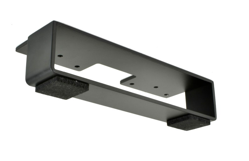 May include: A black metal bracket with a rectangular shape, featuring a cut-out section and several screw holes. Two dark gray foam pads are attached to the bottom. The bracket is designed for mounting.