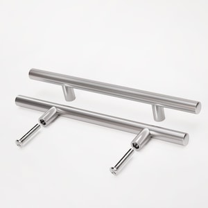 Puede incluir: Dos tiradores de armario de níquel cepillado con tornillos de montaje. Los tiradores cilíndricos son rectos y modernos, diseñados para un agarre fácil. El herraje se muestra sobre un fondo blanco, resaltando el acabado metálico.