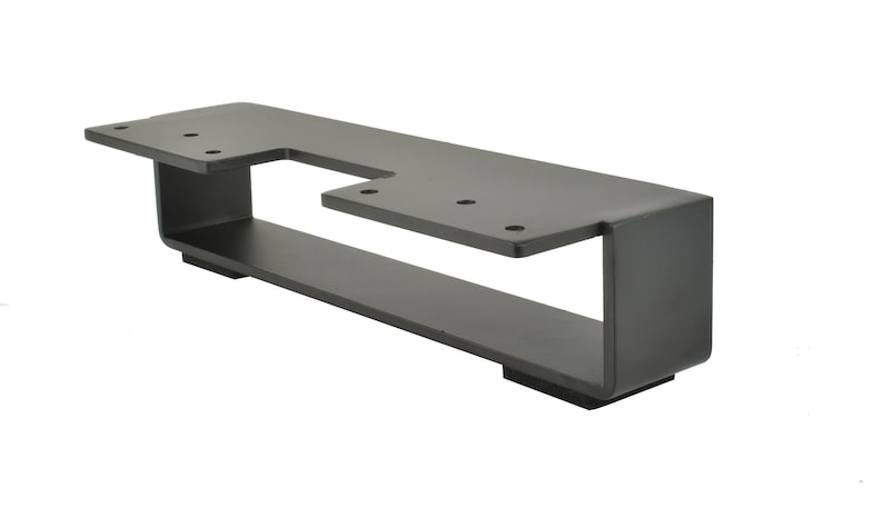 May include: A matte black metal bracket with a unique cut-out design. The bracket has a rectangular base with a raised, angled top section. Several holes are drilled for mounting. The overall design is modern and industrial.