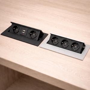 Könnte beinhalten: Zwei Pop-up-Steckdosen, eine schwarze und eine silberne, mit mehreren Steckdosen und USB-Anschlüssen. Konzipiert für den bündigen Einbau in einen Schreibtisch oder Tisch, bietet bequemen Zugang zu Strom und Ladefunktionen. Metrische Maße.