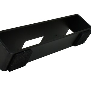 May include: A black rectangular stand with a hollow interior and two dark gray felt pads on the bottom. The stand has a slanted front and two cut-out sections.