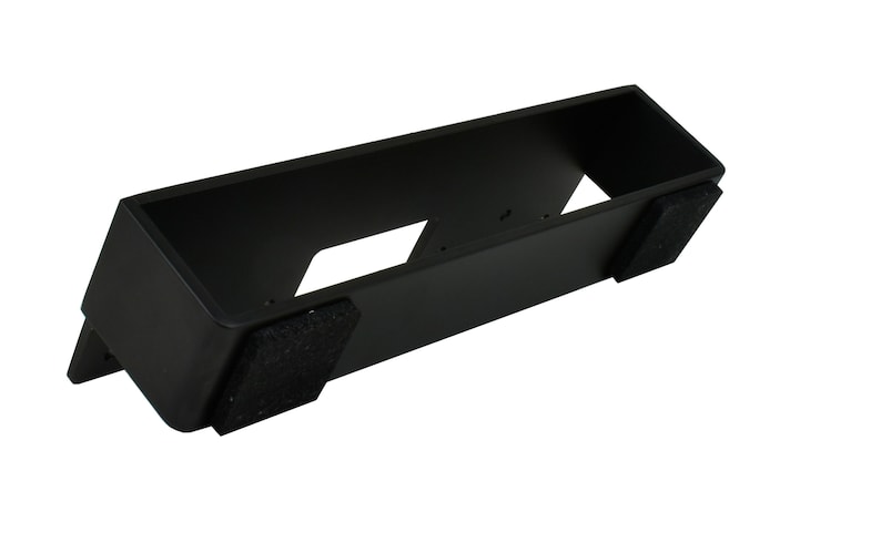 May include: A black rectangular stand with a hollow interior and two dark gray felt pads on the bottom. The stand has a slanted front and two cut-out sections.