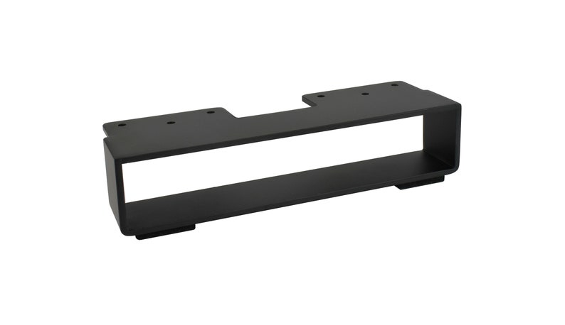 May include: A black metal bracket with a rectangular design. The bracket has a flat top with pre-drilled holes and a lower shelf. The overall shape is a long rectangle with a cutout.