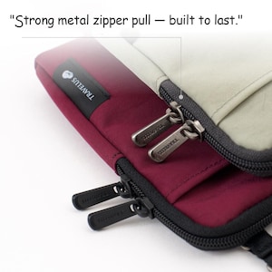 K&ouml;nnte beinhalten: Nahaufnahme eines weinroten und beigefarbenen Reiseaccessoires mit einem schwarzen Rei&szlig;verschluss. Die Metall-Rei&szlig;verschlussanh&auml;nger sind mit "TRAVELUS" graviert. Der Text "Strong metal zipper pull - built to last." ist sichtbar.