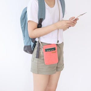 Puede incluir: Una bandolera de color coral con un acento verde y blanco se lleva alrededor del cuello. La bolsa es rectangular y tiene un peque&ntilde;o bolsillo. Una persona lleva la bolsa, una camiseta blanca, pantalones cortos caqui y una mochila azul.