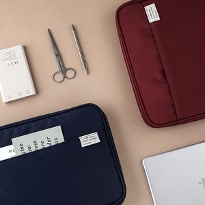 May include: Two zippered laptop sleeves, one navy blue and one burgundy, with a front pocket and a small label that says "A Side Will", both on a beige surface.