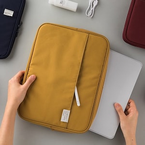 Puede incluir: Una funda para portátil de color mostaza con un bolsillo con cremallera en la parte delantera. La funda está abierta y un portátil blanco está en el interior. La funda está hecha de un tejido suave y duradero.