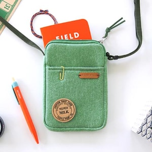 Puede incluir: Un bolso cruzado verde con un peque&ntilde;o bolsillo delantero, un clip dorado y un parche de cuero marr&oacute;n. Dentro hay un cuaderno naranja con la palabra "FIELD" impresa. Otros art&iacute;culos incluyen un bol&iacute;grafo, una lata y una tarjeta de bingo.