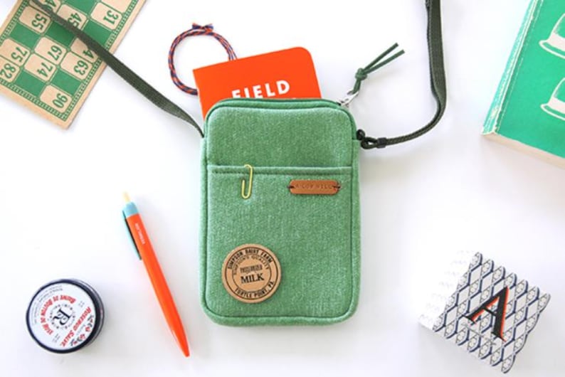 Puede incluir: Un bolso cruzado verde con un peque&ntilde;o bolsillo delantero, un clip dorado y un parche de cuero marr&oacute;n. Dentro hay un cuaderno naranja con la palabra "FIELD" impresa. Otros art&iacute;culos incluyen un bol&iacute;grafo, una lata y una tarjeta de bingo.
