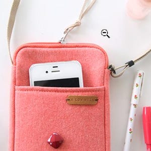 Puede incluir: Un bolso cruzado para tel&eacute;fono de color coral con un tel&eacute;fono inteligente blanco en el bolsillo delantero. El bolso tiene una correa beige y una peque&ntilde;a etiqueta de cuero marr&oacute;n que dice "A LOW HILL". Una gema roja est&aacute; en la parte inferior de la bolsa.
