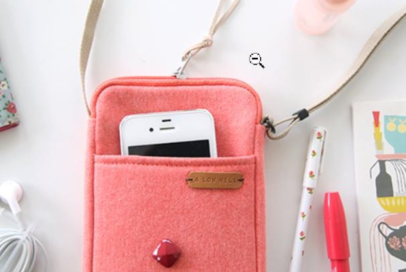 Puede incluir: Un bolso cruzado para tel&eacute;fono de color coral con un tel&eacute;fono inteligente blanco en el bolsillo delantero. El bolso tiene una correa beige y una peque&ntilde;a etiqueta de cuero marr&oacute;n que dice "A LOW HILL". Una gema roja est&aacute; en la parte inferior de la bolsa.